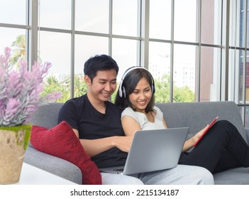 Young Asian Couple Wear Casual Clothing Relax Sitting On Cozy Couch In Living Room. Man Working On Laptop. Woman With Headphones And Playing With Tablet PC, Work At Home Concept. WFH