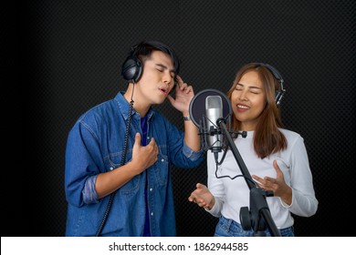 Young Asian Couples With Headphones Sing In Chorus In Front Of Black Soundproofing Walls. Musicians Producing Music In Professional Recording Studio.