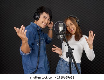 Young Asian Couples With Headphones Sing In Chorus With Microphone And Pop Filter On Tripod Stand. Musicians Producing Music In Professional Recording Studio.