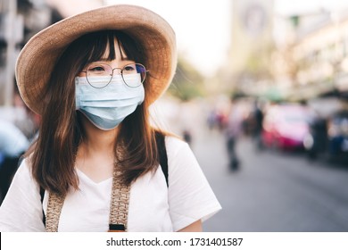 Young Asian Cute Traveller Glasses Girl Wear Mask Protect PM 2.5 And Virus COVID-19. College Student Backpack Travel City With New Normal Lifestyle. Blur Background With Copy Space.