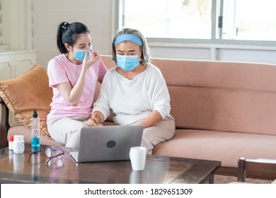 Young Asian Daughter Wearing Surgical Face Mask To Her Mother Sitting In Living Room At Home. Online Learning On Laptop Computer. Protect Against Coronavirus