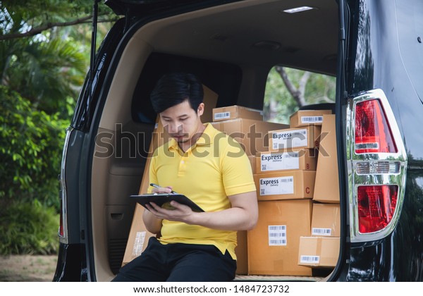 Young Asian Delivery man in yellow uniform\
checking list of package boxes on van before delivering to\
customer, Delivery\
Service