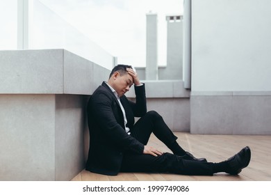 Young Asian Depressed Man Sitting On Outdoors Street Near Office Business Center Building. The Upset Male Businessman Lost His Job Due To The Financial Crisis And Quarantine. Businessman Has Problems