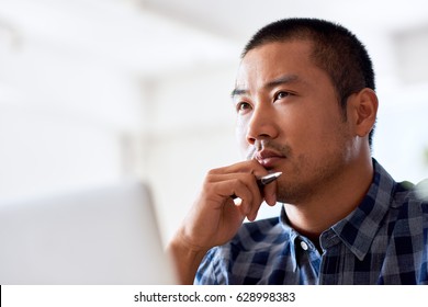Young Asian designer deep in thought with a hand on his chin while working on a laptop alone at a desk in a modern office