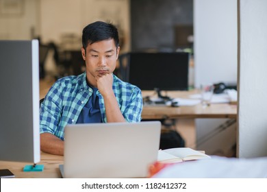 Young Asian designer focused on his work while sitting at his desk in a modern office working online with a laptop - Powered by Shutterstock