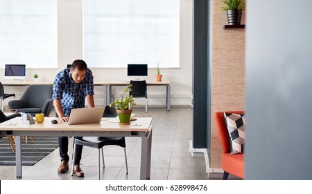 Young Asian designer focused on working on a laptop while leaning on his desk in a bright modern office