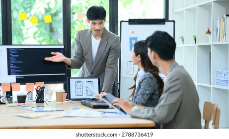 Young Asian developer presents mobile app UI UX wireframes and code to colleagues in a collaborative office brainstorm session, ideal for tech and startup concepts.