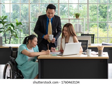 young asian disabled woman with smiling face sitting in the wheelchair and using computer to discuss project with her colleagues in the working office. disability and handicapped concept