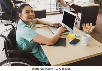 Young Asian Disabled Woman With Smiling Face Sitting In The Wheelchair And Using Computer To Discuss Project With Her Colleagues In The Working Office. Disability And Handicapped Concept