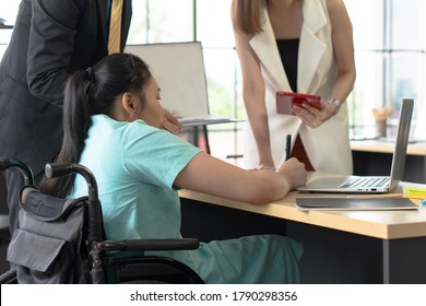 Young Asian Disabled Woman With Smiling Face Sitting In The Wheelchair And Using Computer To Discuss Project With Her Colleagues In The Working Office. Disability And Handicapped Concept