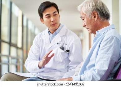 Young Asian Doctor Talking And Explaining Test Result And Diagnosis To Demoralized Elderly Patient In Hospital Hallway