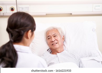 Young Asian Doctor Talking To Senior Patient At Bedside In Hospital Ward