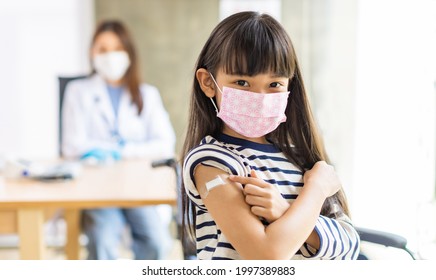 Young Asian Doctor Wearing Gloves And Isolation Mask Is Making A COVID-19 Vaccination In The Shoulder Of Child Patient  At Hospital.