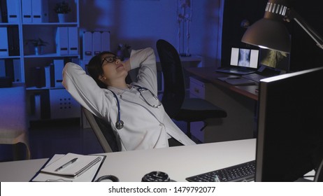 Young Asian Doctor Woman Sitting At Dark Clinic Office In Chair And Reclining On Back. Medical Nurse Smiling While With Relaxation Looking At Ceiling In Hospital At Night. Stay Up Late Working.