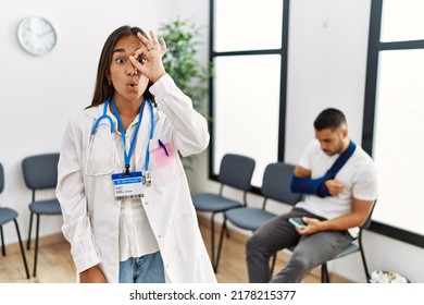 Young Asian Doctor Woman At Waiting Room With A Man With A Broken Arm Doing Ok Gesture Shocked With Surprised Face, Eye Looking Through Fingers. Unbelieving Expression. 