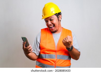 Young asian engineer worker wearing orange vest and helmet over isolated white background celebrating surprised while looking at his phone, amazed for success with left arm raised