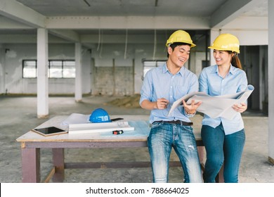 Young Asian Engineers Couple Working Together On Building Blueprint At Construction Site Or Factory. Civil Engineering, Industrial Business Partner, Or Home Renovation Service Concept. With Copy Space