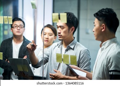 Young Asian Entrepreneur Of Small Company Drawing A Diagram On Glass During Team Meeting Discussing And Analyzing Business Situation In Office.