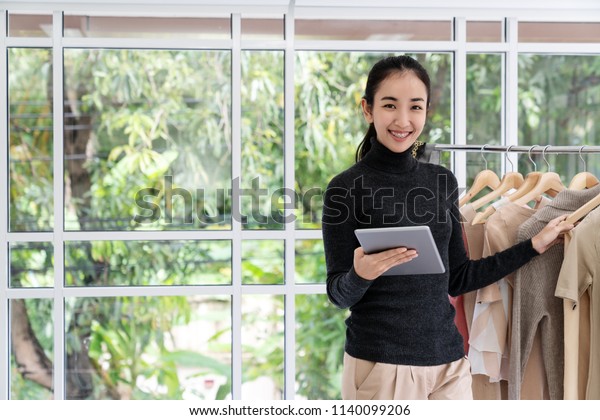 Young asian entrepreneur woman using tablet work\
at home office looking at camera with happy casual lifestyle.\
Portrait of female business owner or fashion designer holding\
smartphone or gadget.