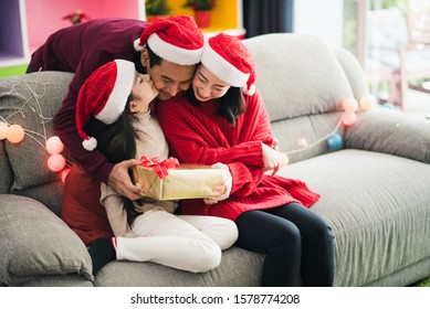 Young Asian Family Celebration In Christmas Day, Parent And Daughter Siting On Sofa Which Hold Give Box, Girl Kiss Father Which Smiling Felling Happy In Living Room At Home Morning Time. Merry Xmas.