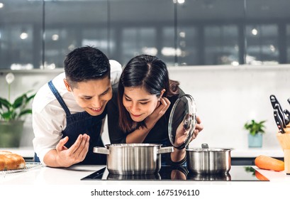 Young Asian Family Couple Having Fun Standing Near Stove And Cooking Together.Happy Couple Looking And Smelling Tasting  Fresh Delicious From Soup In A Pot With Steam At White Interior Kitchen
