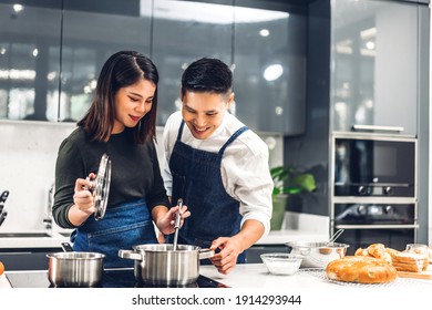 Young Asian Family Couple Having Fun Standing Near Stove And Cooking Together.Happy Couple Looking And Smelling Tasting Fresh Delicious From Soup In A Pot With Steam At White Interior Kitchen