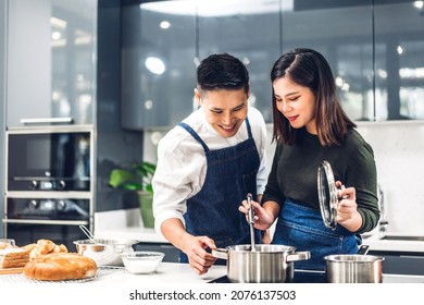 Young Asian Family Couple Having Fun Standing Near Stove And Cooking Together.Happy Couple Looking And Smelling Tasting Fresh Delicious From Soup In A Pot With Steam At White Interior Kitchen