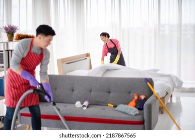 Young Asian Family, The Husband And Wife Are Cleaning The House Together. Man Is Wearing A Red Apron, He Is Vacuuming Using A Vacuum Cleaner, And The Wife Is Wearing The Same Apron