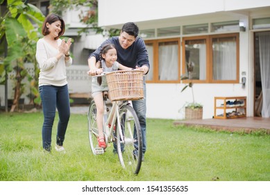 Young Asian Family Love, Father And Mother Teach And Training Daughter To Ride A Bike On Grass Field At Outdoor Of Home. Girl Train To Ride A Bike By Her Parent Which Smiling And Felling Happy