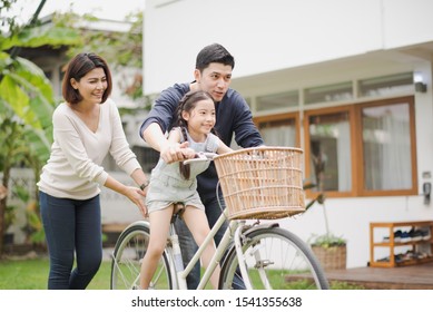 Young Asian Family Love, Father And Mother Teach And Training Daughter To Ride A Bike On Grass Field At Outdoor Of Home. Girl Train To Ride A Bike By Her Parent Which Smiling And Felling Happy