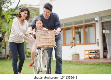 Young Asian Family Love, Father And Mother Teach And Training Daughter To Ride A Bike On Grass Field At Outdoor Of Home. Girl Train To Ride A Bike By Her Parent Which Smiling And Felling Happy