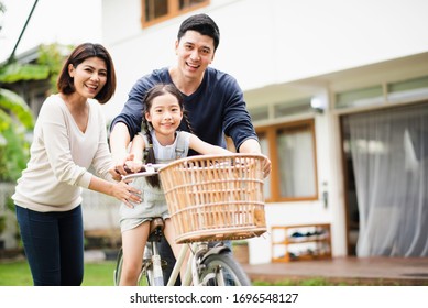 Young Asian family love, father and mother teach and training daughter to ride a bike on grass field at outdoor of home. girl train to ride a bike by her parent which smiling and felling happy