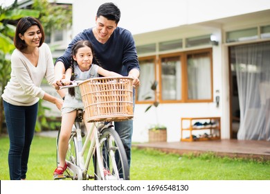 Young Asian Family Love, Father And Mother Teach And Training Daughter To Ride A Bike On Grass Field At Outdoor Of Home. Girl Train To Ride A Bike By Her Parent Which Smiling And Felling Happy