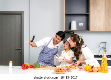 Young Asian Family Are Preparing Salad In The Kitchen And Father Take A Photo Selfie By Phone. Excited Smiling And Felling Happy. Parent Teach Daughter To Cooking Food On The Day At Home.
