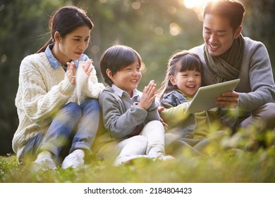 Young Asian Family With Two Children Sitting On Grass Outdoors In Park Watching Contents In Tablet Computer