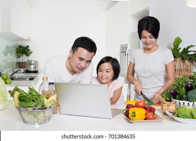 Young Asian Family Using A Laptop While Preparing Meal.