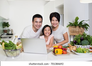 Young Asian Family Using A Laptop While Preparing Meal.