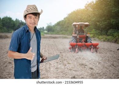 Young Asian Farmer Holding A Tablet In A Field With A Tractor Adjusting The Soil