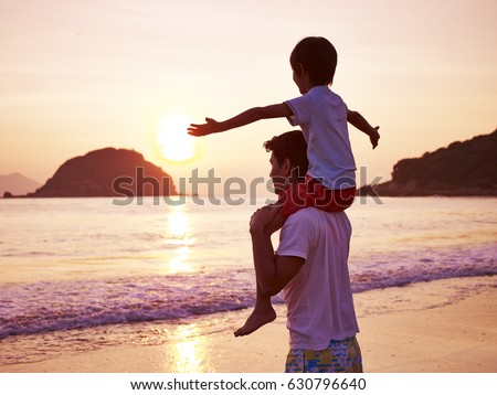 young asian father carrying son on shoulder on beach at sunrise.