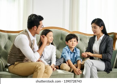 Young Asian Father And Mother And Two Children Sitting On Couch Chatting In Family Living Room At Home