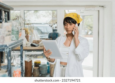 Young asian female artist in headscarf talking on smartphone and using digital tablet while working in blurred ceramic workshop at background, creative process of pottery making - Powered by Shutterstock