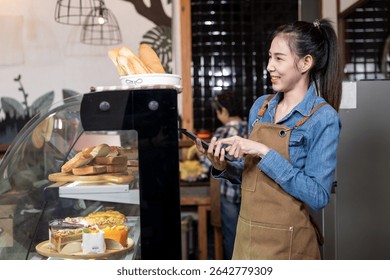 Young Asian female barista in denim shirt and brown apron holding digital tablet while standing near bakery display filled with assorted breads and cakes. Smiling warmly while managing orders in cafe