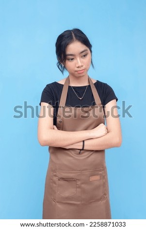 A young asian female barista feeling down and sad. Wearing a brown apron and black shirt and against a light blue background.