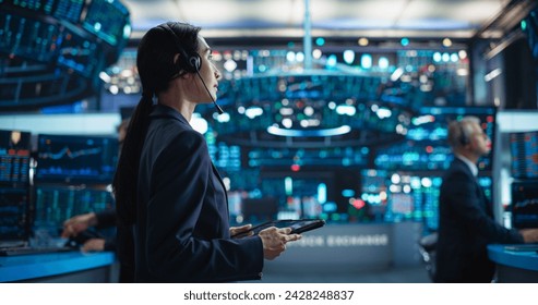 Young Asian Female Broker Working on Tablet Computer on Stock Exchange Floor. Traders Monitoring Real-Time Stocks, Commodities and Exchange Market Data. Professional Investment Agents in Office - Powered by Shutterstock