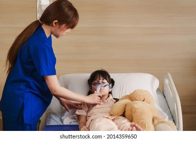 Young Asian Female Caregiver Holding Oxygen Mask With Cute Girl Patient In Hospital. Nurse Putting Putting Inhalation On The Girl Face.