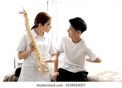 A young Asian female chiropractor instructs a young Asian male patient using a spinal model. - Powered by Shutterstock