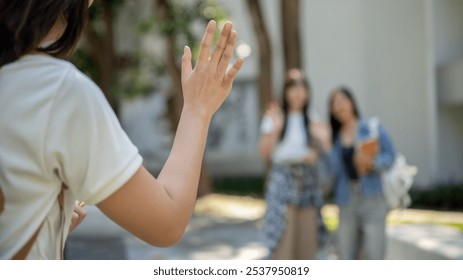 A young Asian female college student is waving to her friends, saying hello or goodbye, while passing by in her college. a close-up image with a copy space. education and friendship concepts