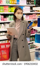 Young Asian Female Consumer With Red Basket And Tablet Standing By Large Shelf With Haircare Products And Looking Through Their Characteristics