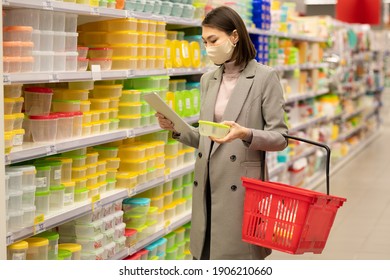 Young Asian Female Consumer With Red Shopping Basket Standing By Large Shelf With Containers And Looking Through Their Characteristics In Tablet