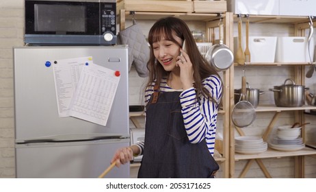 Young Asian Female Cooking In Kitchen Having Pleasant Phone Talk. Taiwanese Lady Wearing Apron Walking Away From Stove With Wooden Spoon In Hand. Burst Out Laughing. Genuine Lifestyle